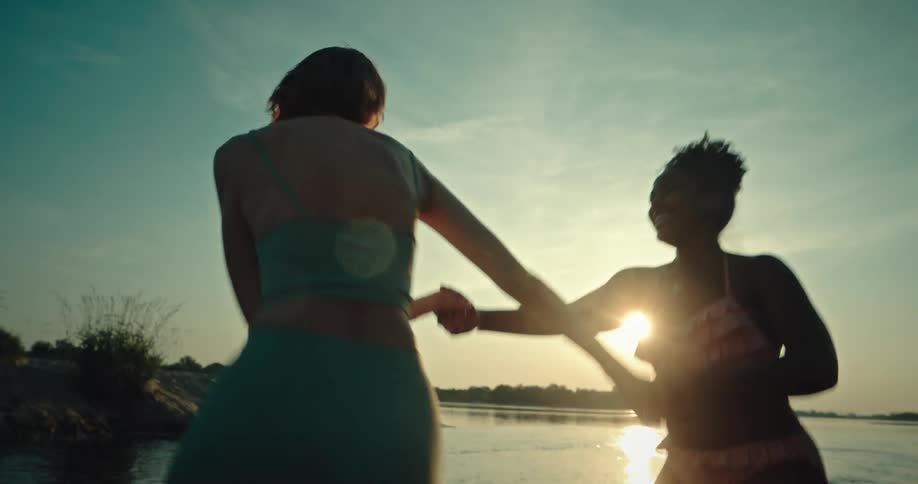 Two women wearing bikinis and holding hands run in the water at sunset