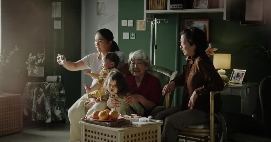 Grandmother sitting in an armchair with her granddaughters and daughters beside her taking a selfie in a hospital room