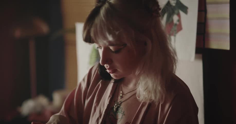 Young female with two toned hair retrieves sewing pins from a small box in a room dim lit with natural sunlight with clothing design drawings on the wall in the background