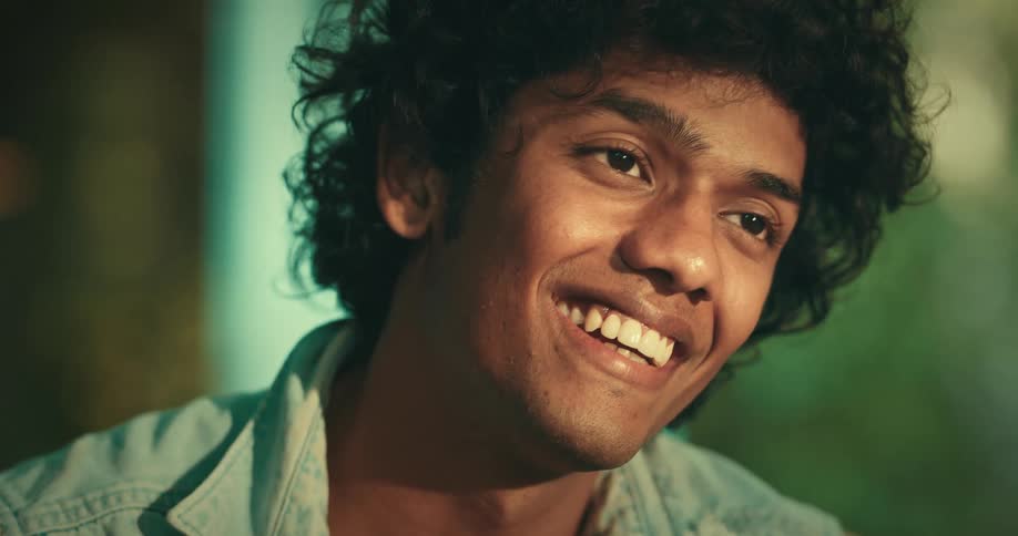A young man with curly hair smiles while he is talking to someone
