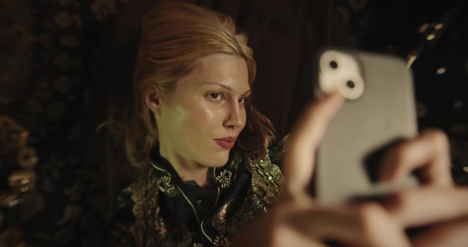 A young blonde woman is lying down and making faces smiling while taking photos of herself with a cell phone