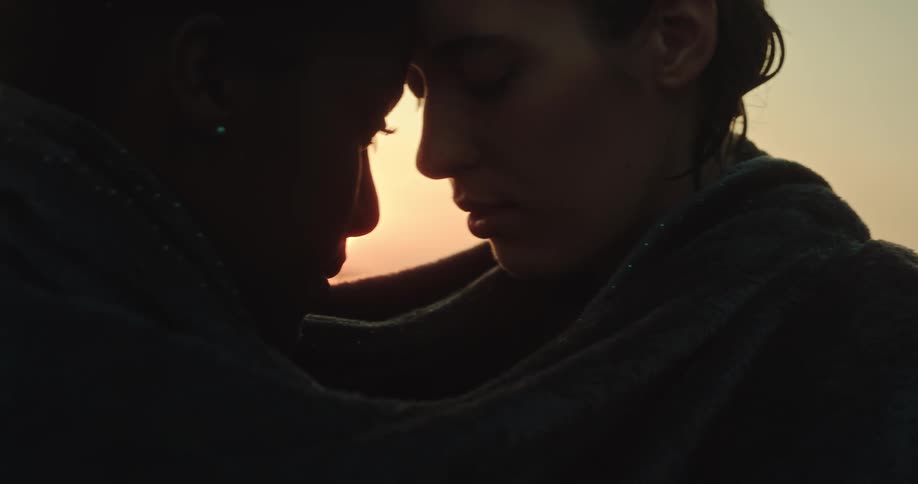 Two women touching foreheads with a towel wrapped around them with the sunset on the horizon