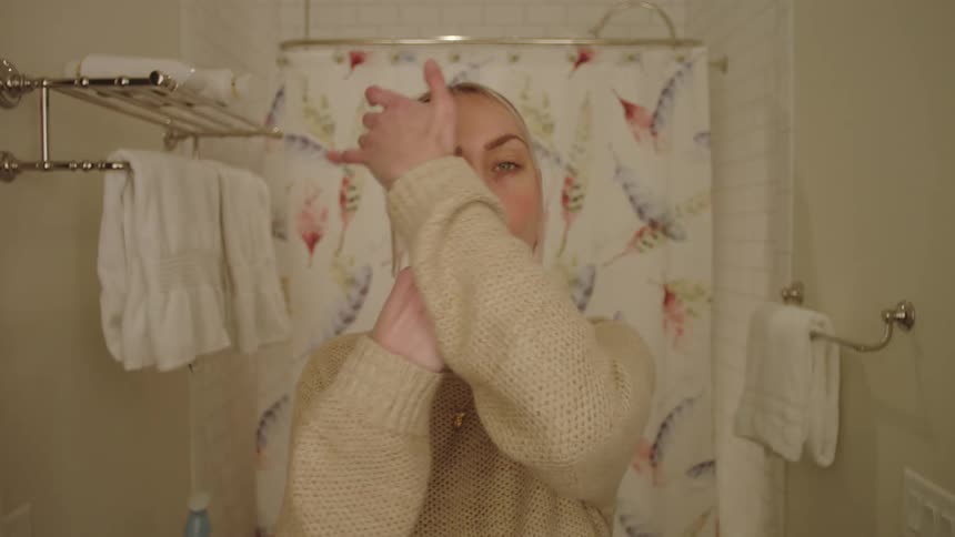 A woman in a small bathroom brushes her teeth and smooths her hair
