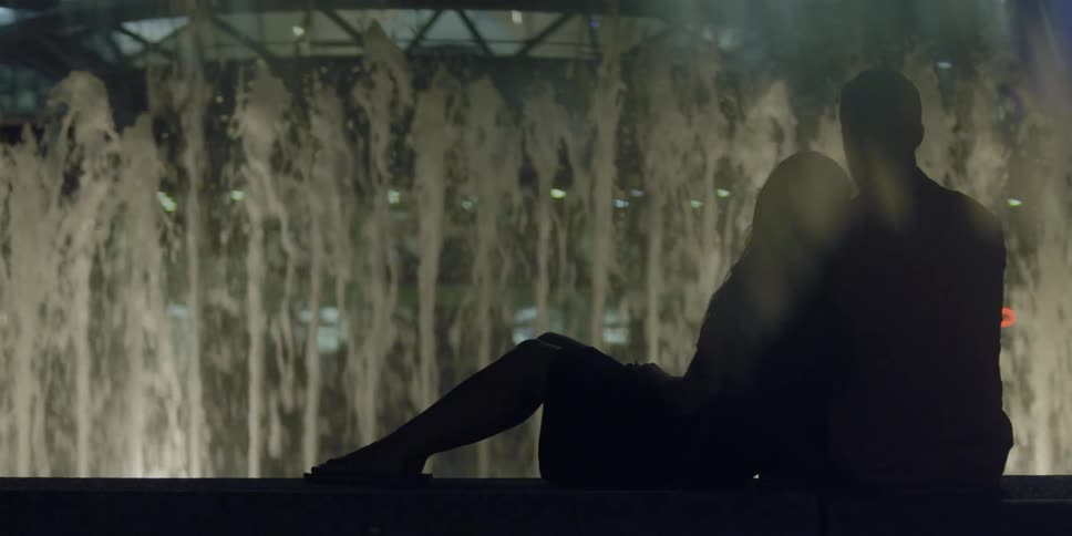 A man and woman embrace near a city fountain at night