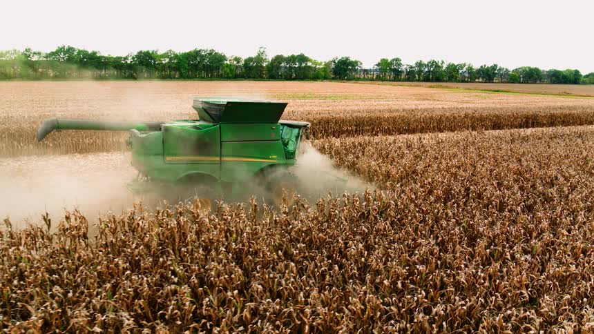 Green tractor harvests crops during the daytime with dust kicking up in the field