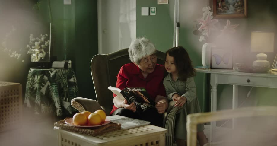 An elderly woman reads to a female child while seated on a chair indoor during the day