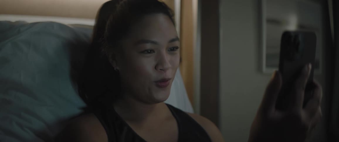 Woman sitting in bed in dimly lit room smiles while making a video call on her smartphone