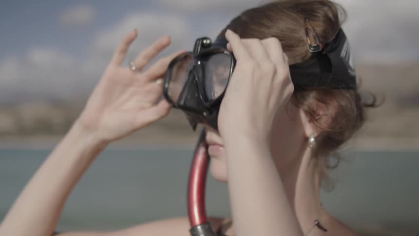 A young woman standing near water lowers her snorkeling mask