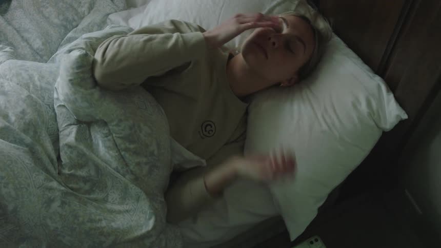 A blonde woman dressed in a brown sweater wakes up and gets off the bed