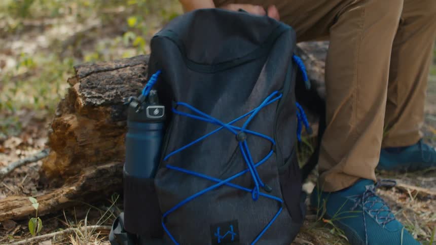 Man put a water bottle by the side of a backpack and lifted the bag up in the woods at midday