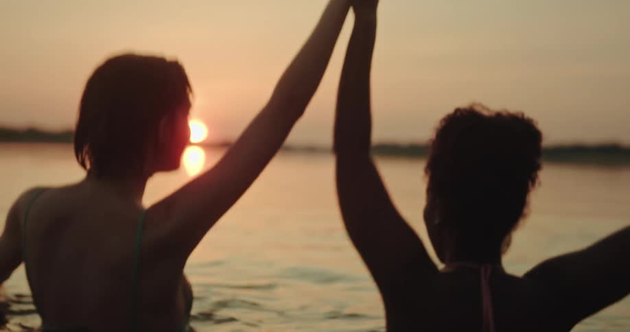 Two women wearing bikinis dance in the water at sunset