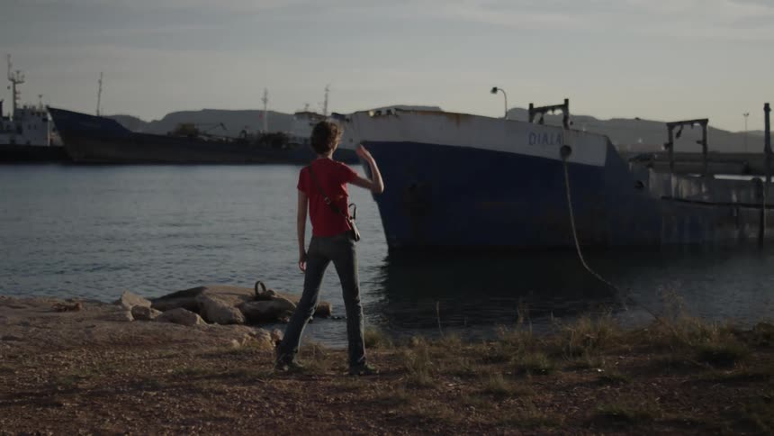 Woman walks along shore across from mountains looking at ships