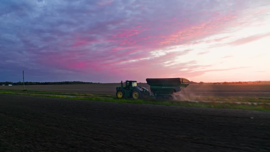 Commercial tractor transporting a grain bin down a country road between two plowed fields at sunset with blue purple and pink clouds in the sky