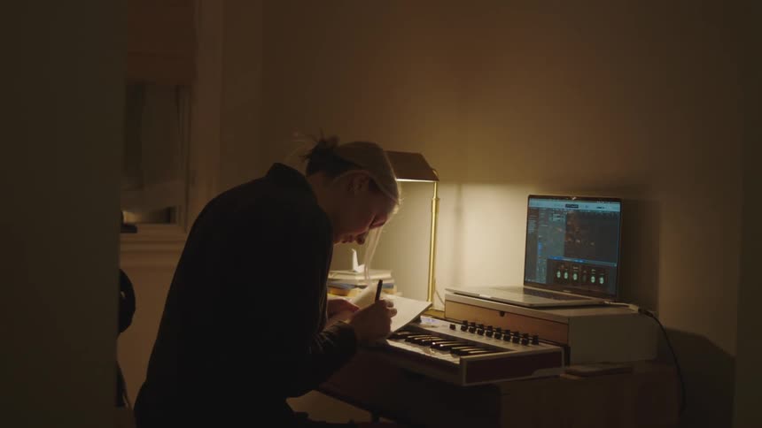 Woman sitting playing a digital piano with a laptop in front of her and writing something in a notebook