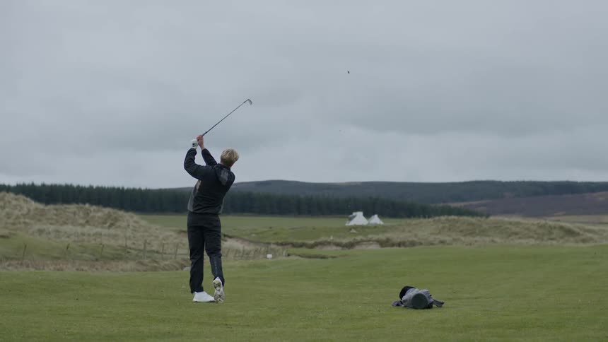 Blonde man wearing back jacket and pants plays golf on a cloudy day