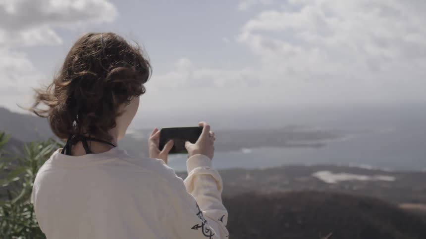 Woman with her back to the camera holding a cellphone taking a photo at the peak of a hill overlooking the sea