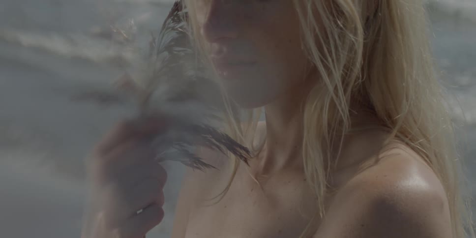 A blond haired woman holds a plant to her face as waves crash behind her