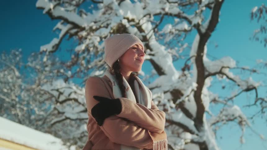 Woman wearing beanie coat and gloves stands in front of snow covered tree