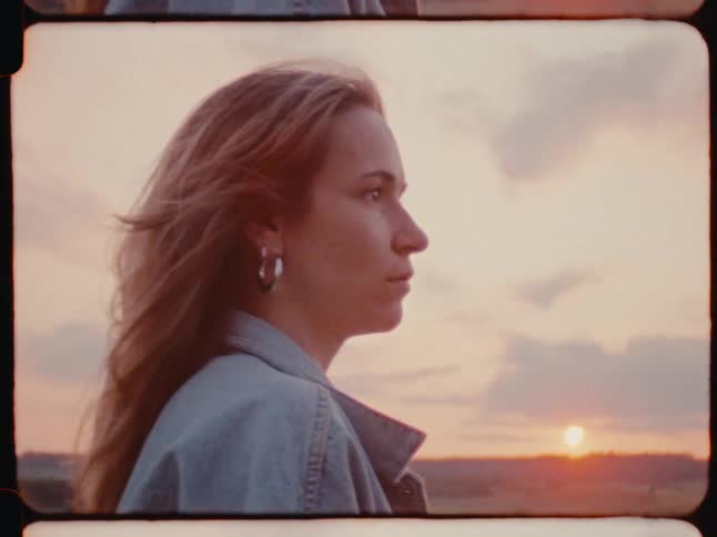 A young woman stands outside in a grassland at sunset