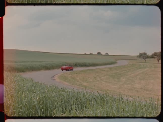 Person drives red convertible past field of crops on a cloudy day