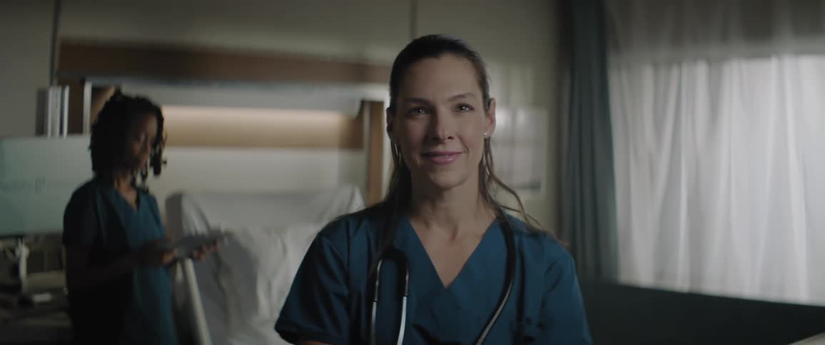 Medical professional stands and smiles while another nurse standing behind her in hospital room uses tablet