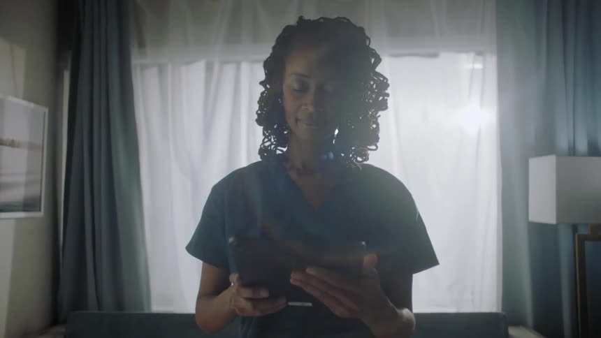 Nurse standing looking at a tablet and smiling in a hospital room in front of a window with sunlight shining in around her
