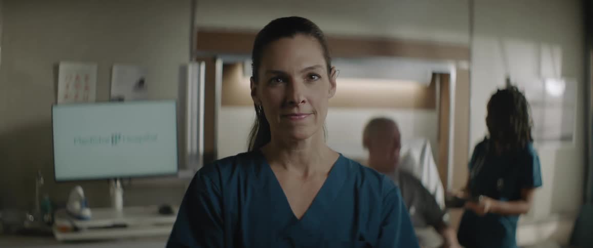 Nurse stands with a smile while another nurse interacts with a hospital patient in the background