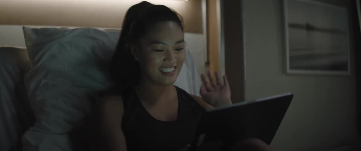 A woman with a ponytail wearing a black undershirt sits on a bed and makes a video call on a tablet