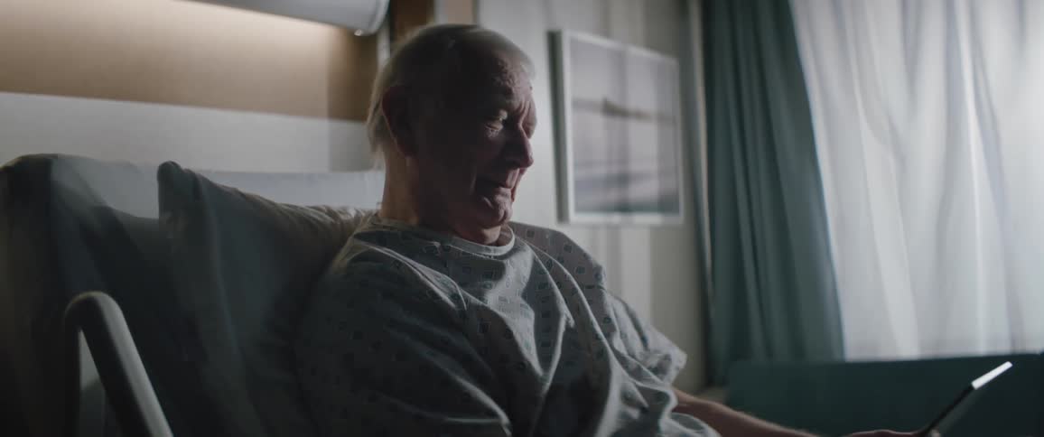 An elderly man on a hospital bed looking at a tablet and smiling at midday