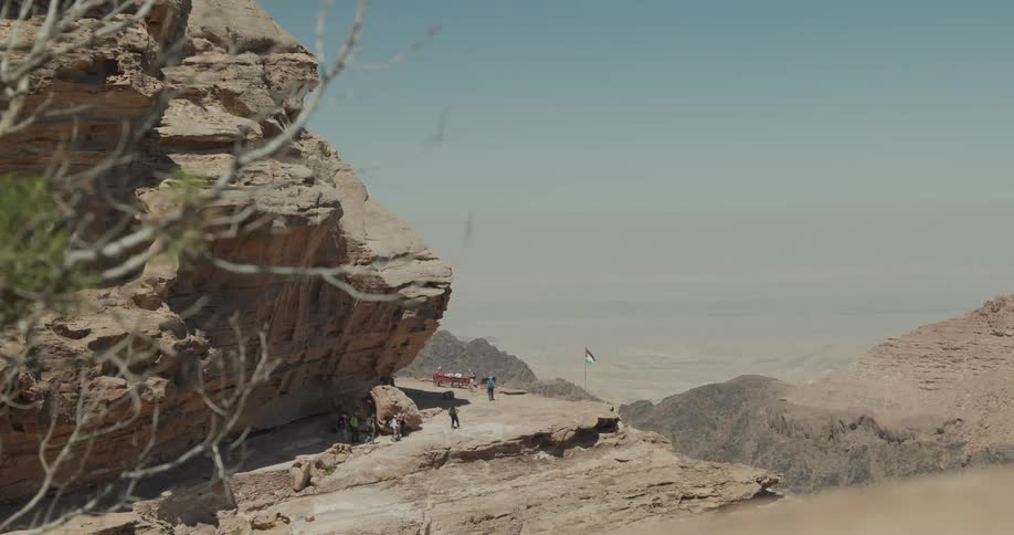 A group of half a dozen hikers walk around the base of a gigantic rock towards an overlook where there are people sitting on a bench