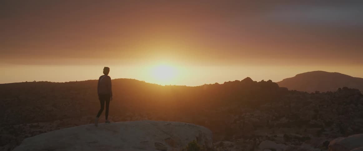 Woman walking on a rock with a panoramic view of a valley and mountains with the sunrise on the horizon