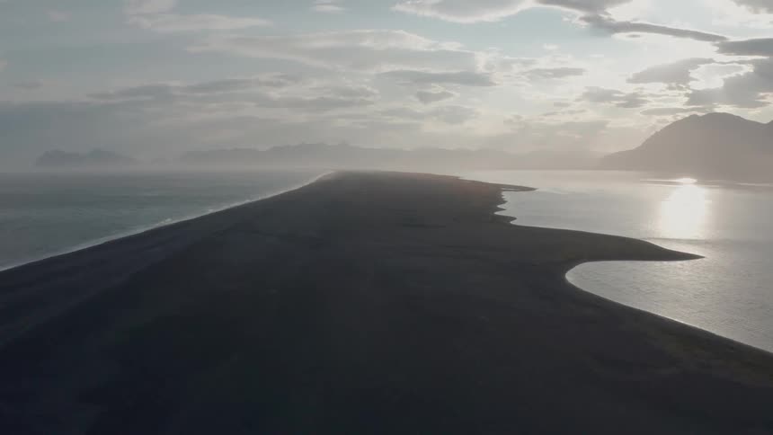 Aerial panorama of a black sand beach next to the sea with a mountain range on the horizon under a morning sky