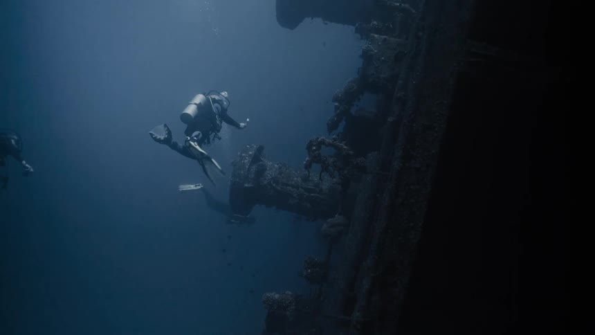 Three divers swim in the ocean amongst coral and artifacts