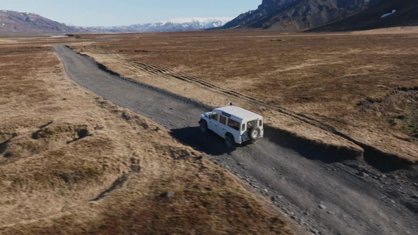 An aerial view of a truck traveling on a dirt road through a wide plain surrounded by rocky mountains during the daytime