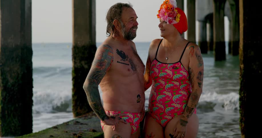 A couple in swimsuits stand below a pier