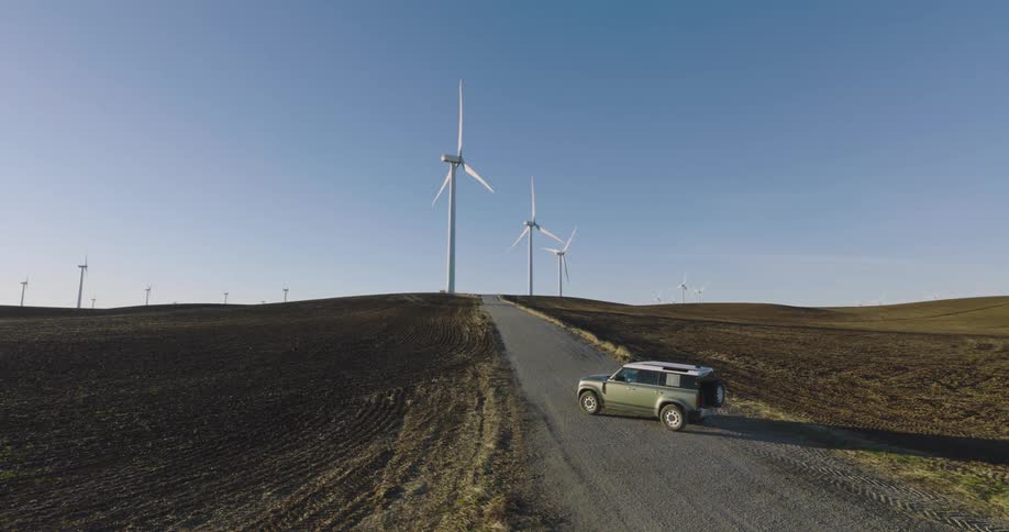 Aerial of a green jeep on dirt road with wind turbines in the background
