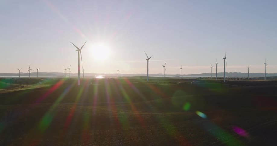 Wind turbines spin on a wind farm as the sun shines brightly in the distance