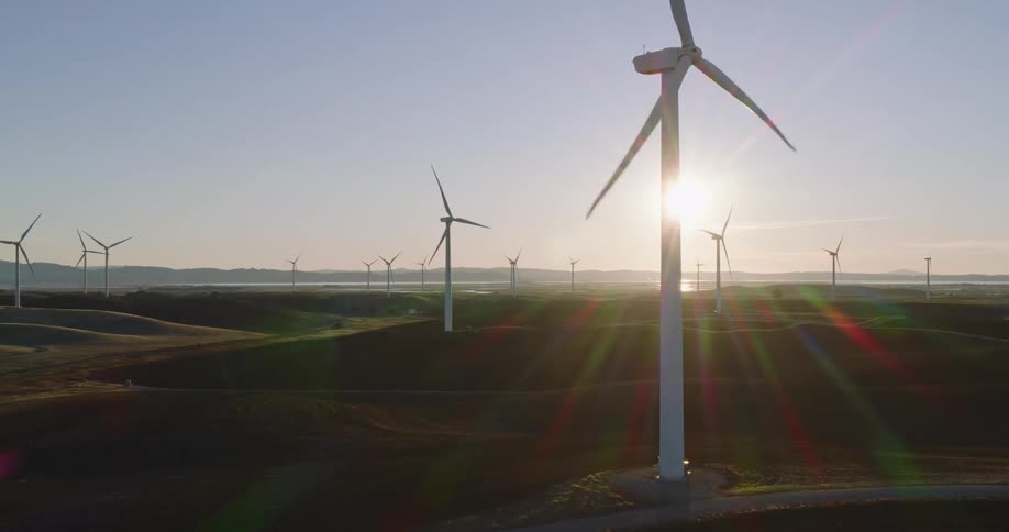 Wind turbines spread out across a grassy countryside on a wind farm at sunset with the sun glaring and clear blue sky in the background