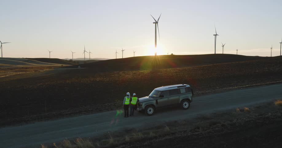 Two workers on an access road by jeep look out to a windfarm with spinning wind turbines at sunset