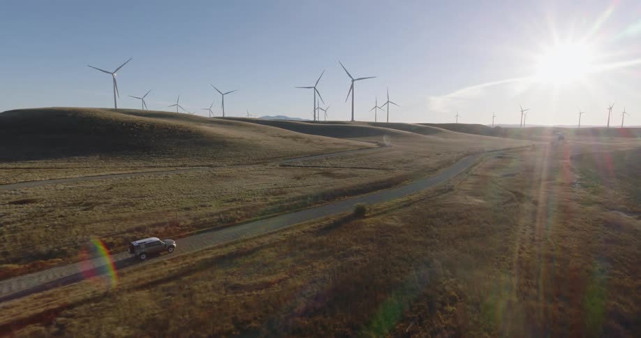 A truck travels on a rural road through a wind farm as turbines spin on the hills