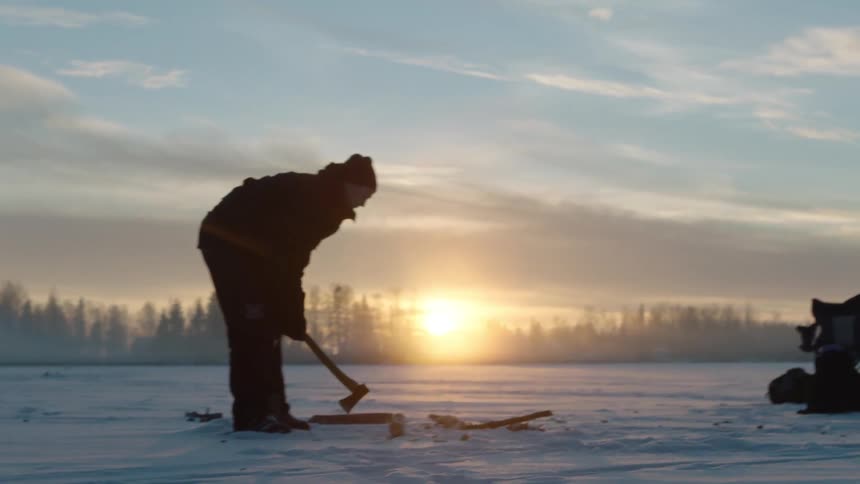 A man is cutting a hole in the ice at sunset