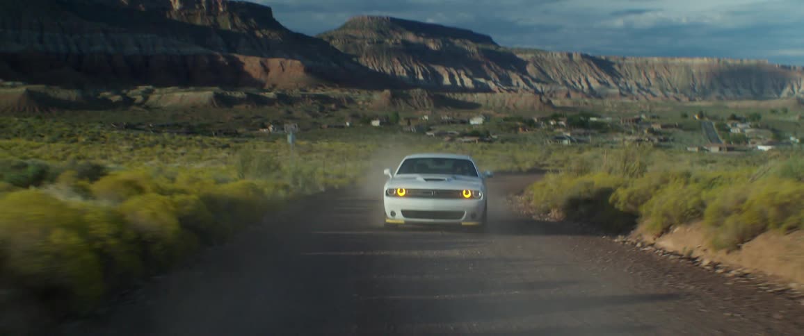 A white sports car is driving on a dirt street in a region with mountains in the background