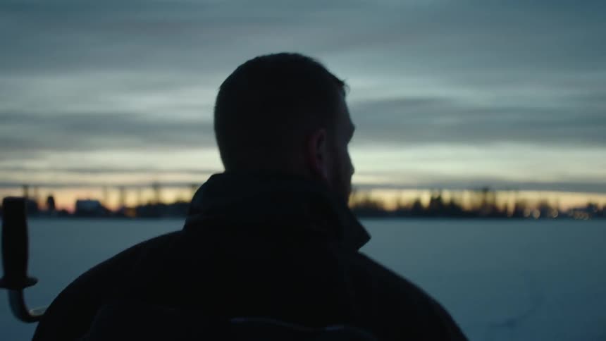 A man carrying a hole digger walks on the ice at dawn