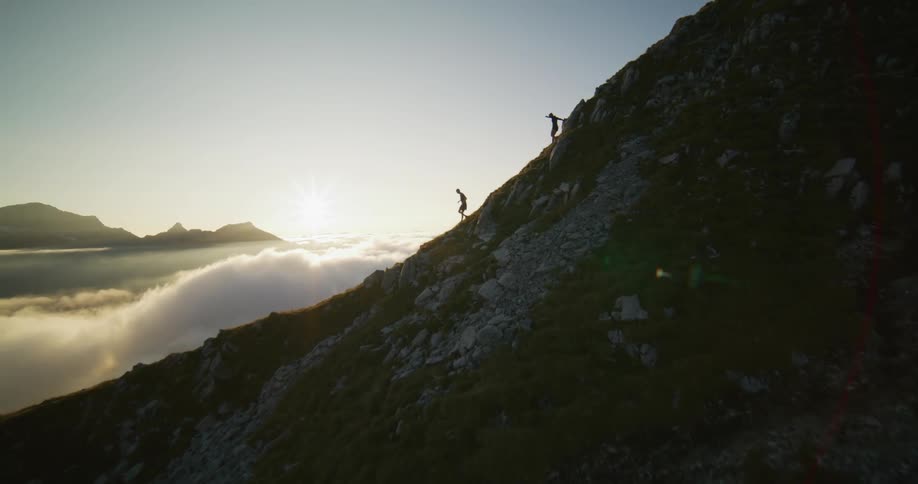 Hikers running down a highland mountain slope ridge surrounded by white clouds at sunset