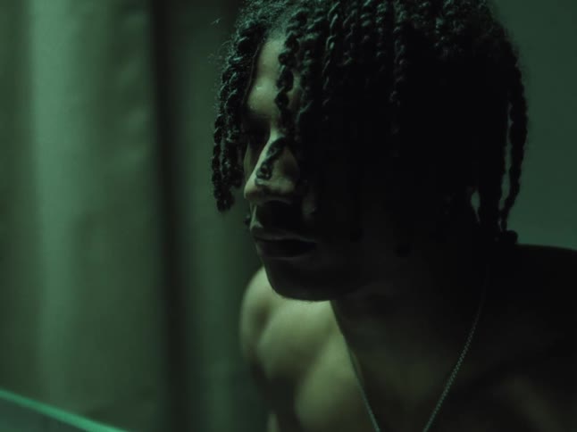 A young black man with braided hair is intently staring at himself in a dimly lit bathroom mirror