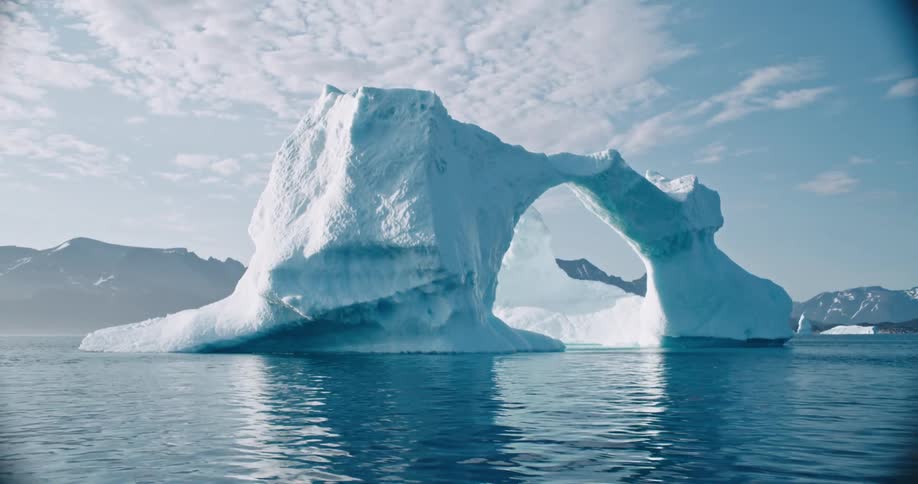 White and blue arch shaped ice formation in a blue sea reflecting a blue and white sky and sunlight on a bright day