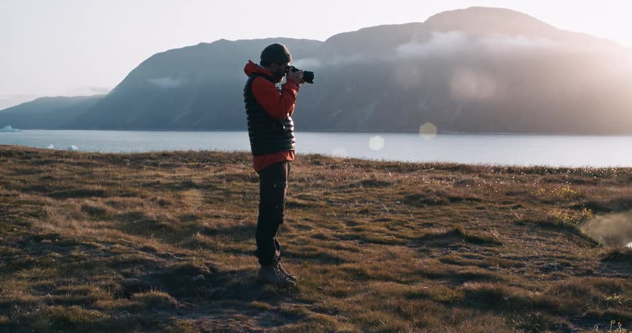 A man in a grassy field taking a photo of a mountain range beyond a lake