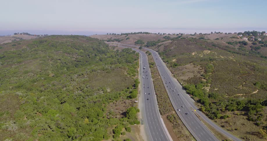 Cars travel along a divided interstate in a valley surrounded by low green hillsides on a sunny day