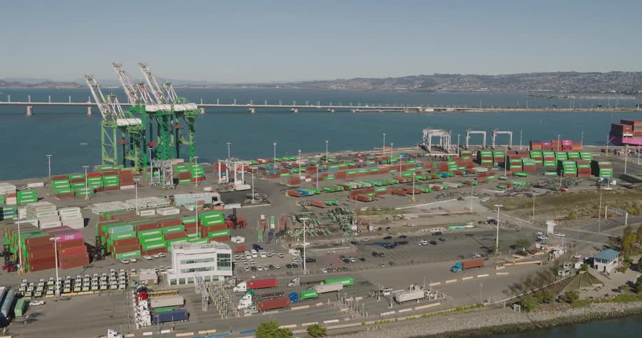 Aerial of shipping container port with large cranes and trucks next to a long bridge on a bright day