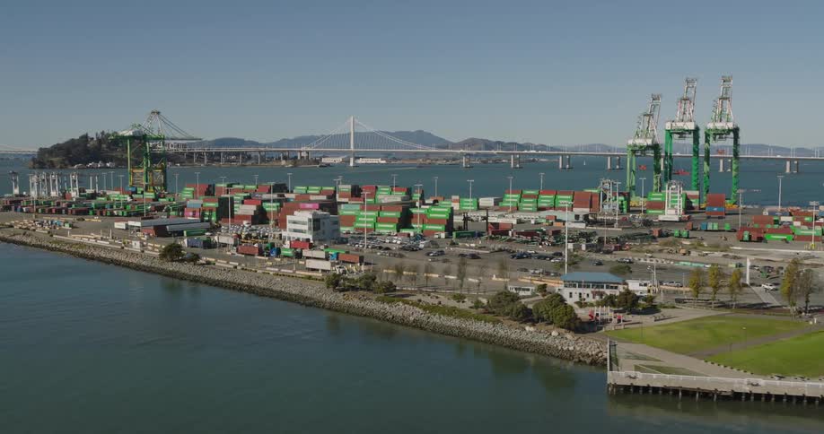 Motor vehicles move along loading dock with mountains and a suspension bridge in the background